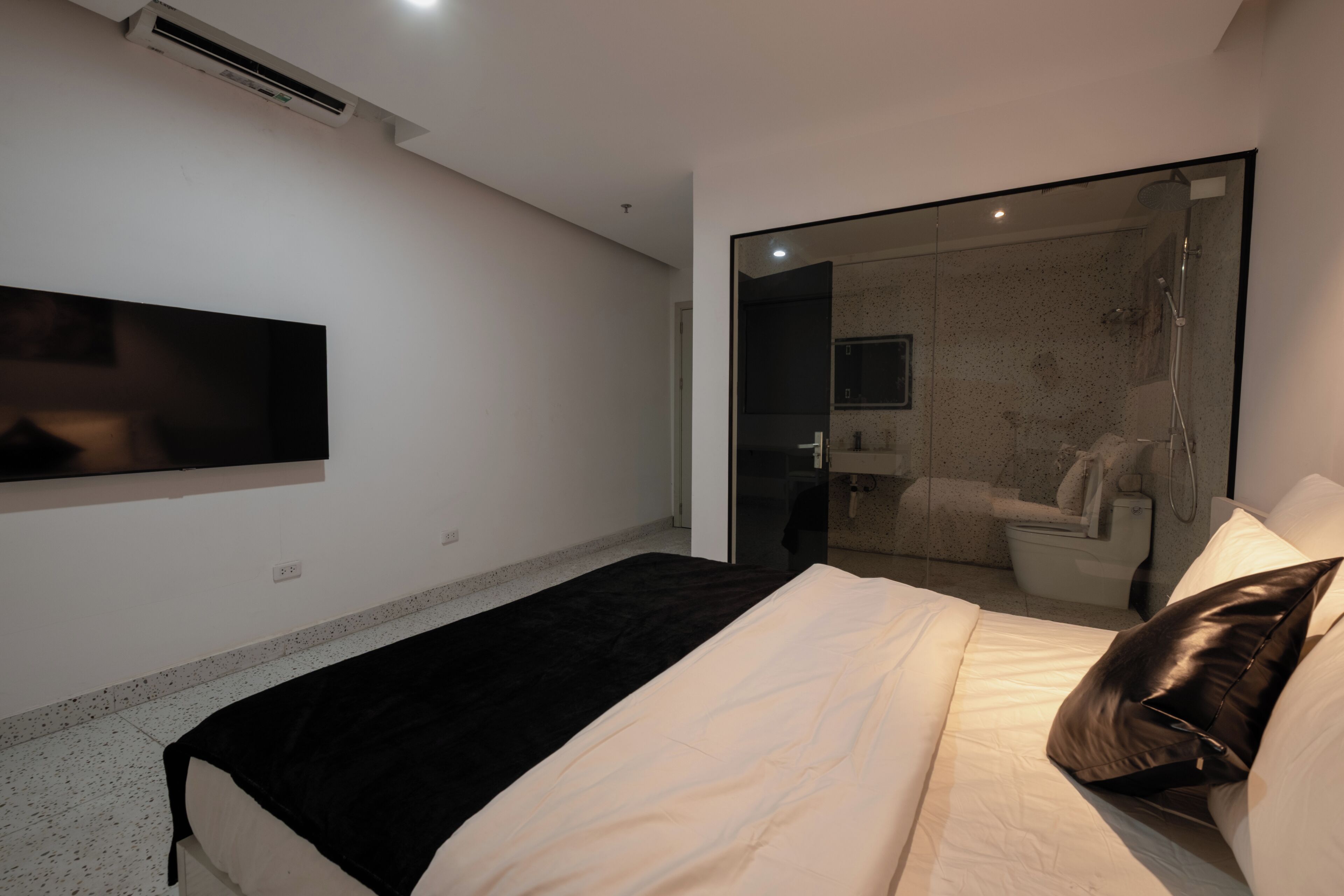 Quarto com cama, manta preta e branca, TV montada e box de vidro.