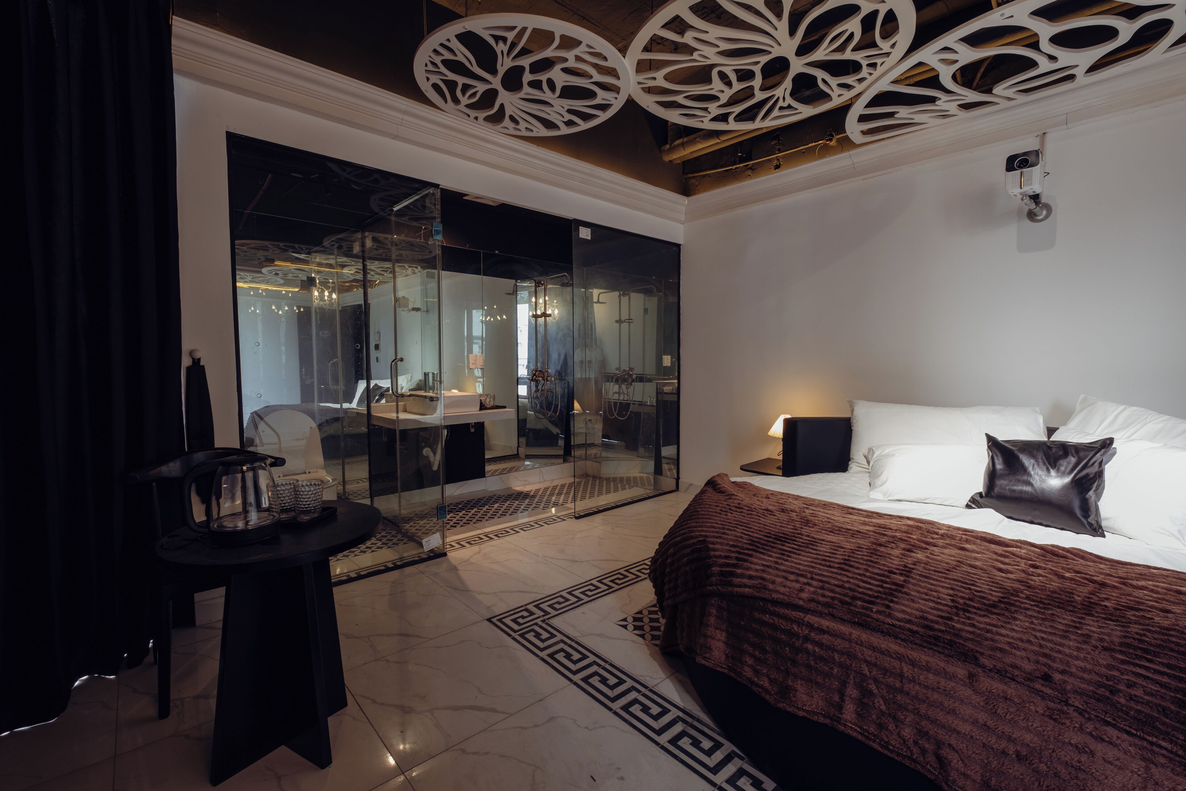 A modern hotel room with a large bed, a glass partition, a dining area, and a chandelier.