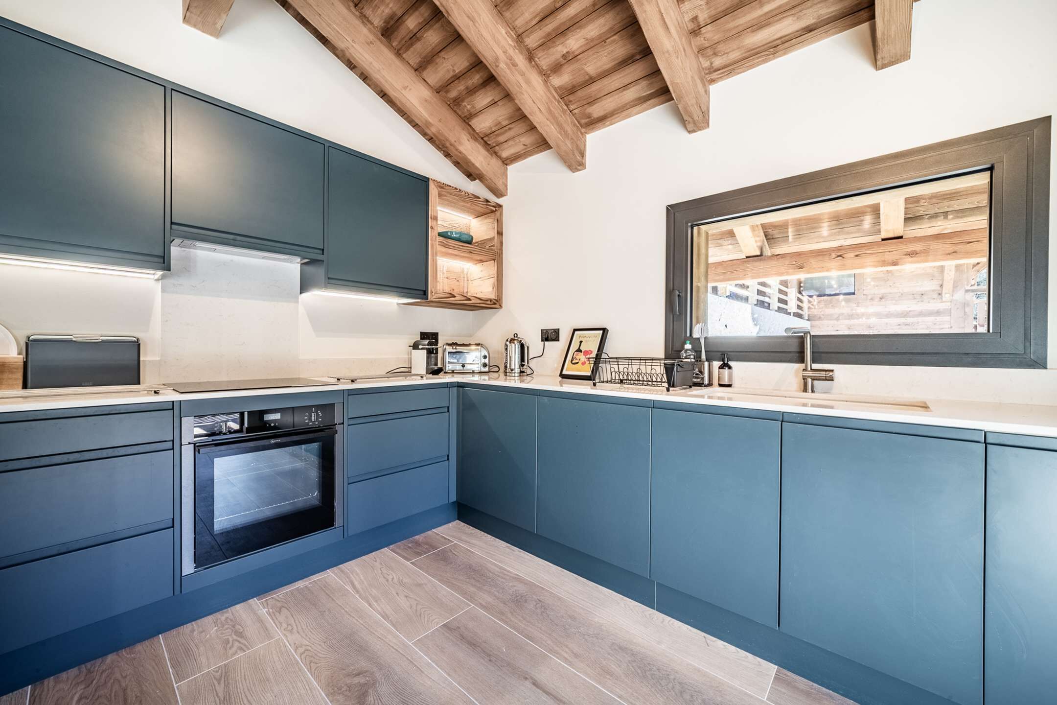 A modern kitchen with blue cabinets, a built-in oven, and a window overlooking a wooden deck.
