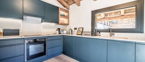A modern kitchen with blue cabinets, a built-in oven, and a window overlooking a wooden deck.
