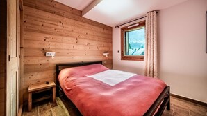 A bedroom with a wooden ceiling, a bed with a red bedspread, a small bedside table, and a window with curtains.