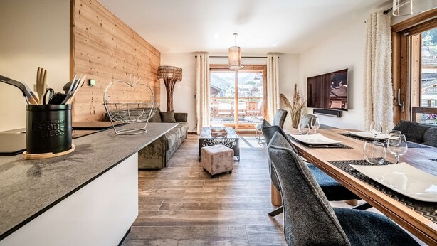 A modern kitchen with a wooden accent wall, a dining area with a long table and chairs, and a view of a mountain landscape through large windows.