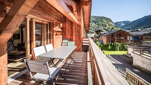 A wooden cabin with a deck, outdoor seating, and a view of a village and mountains.