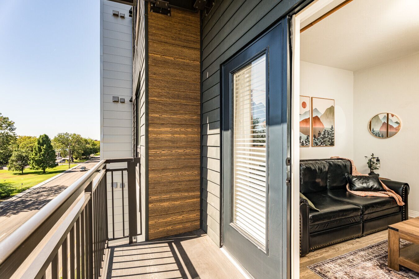 A balcony with a wooden railing, a sliding glass door, and a black leather sofa with a throw blanket.