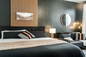A modern bedroom with a large bed, a nightstand, a sofa, a wall-mounted mirror, and a framed picture.