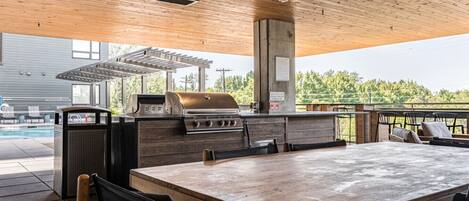 An outdoor patio with a wooden table, chairs, a grill, and a pool in the background.