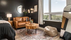 Ein modernes Schlafzimmer mit dunkler Wand, einem braunen Ledersofa, einem runden Holztisch, einem großen Spiegel und Blick ins Grüne durch ein Fenster.