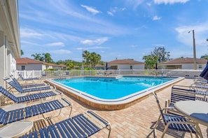 A swimming pool with lounge chairs and tables, surrounded by a fence and palm trees.