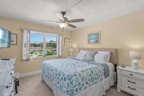 A bedroom with a bed, a ceiling fan, a window with a view, and a nightstand with a lamp.