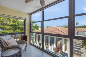 A balcony with a wicker sofa and a small table, large windows offering a view of a residential area with a red-tiled roof. - Modern 2BR | Pool View | Steps to Crescent Beach (Sarasota)