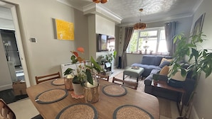 A dining area with a wooden table, chairs, and woven placemats. There is a sofa and a potted plant in the background. - Homely flat in Surrey, Sleeps 3, family friendly (Walton-on-Thames)