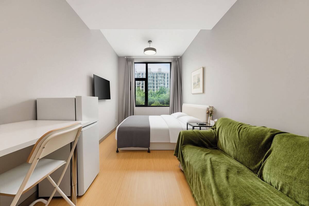 A modern hotel room with a bed, a desk, a green sofa, a TV, and a window with a city view.