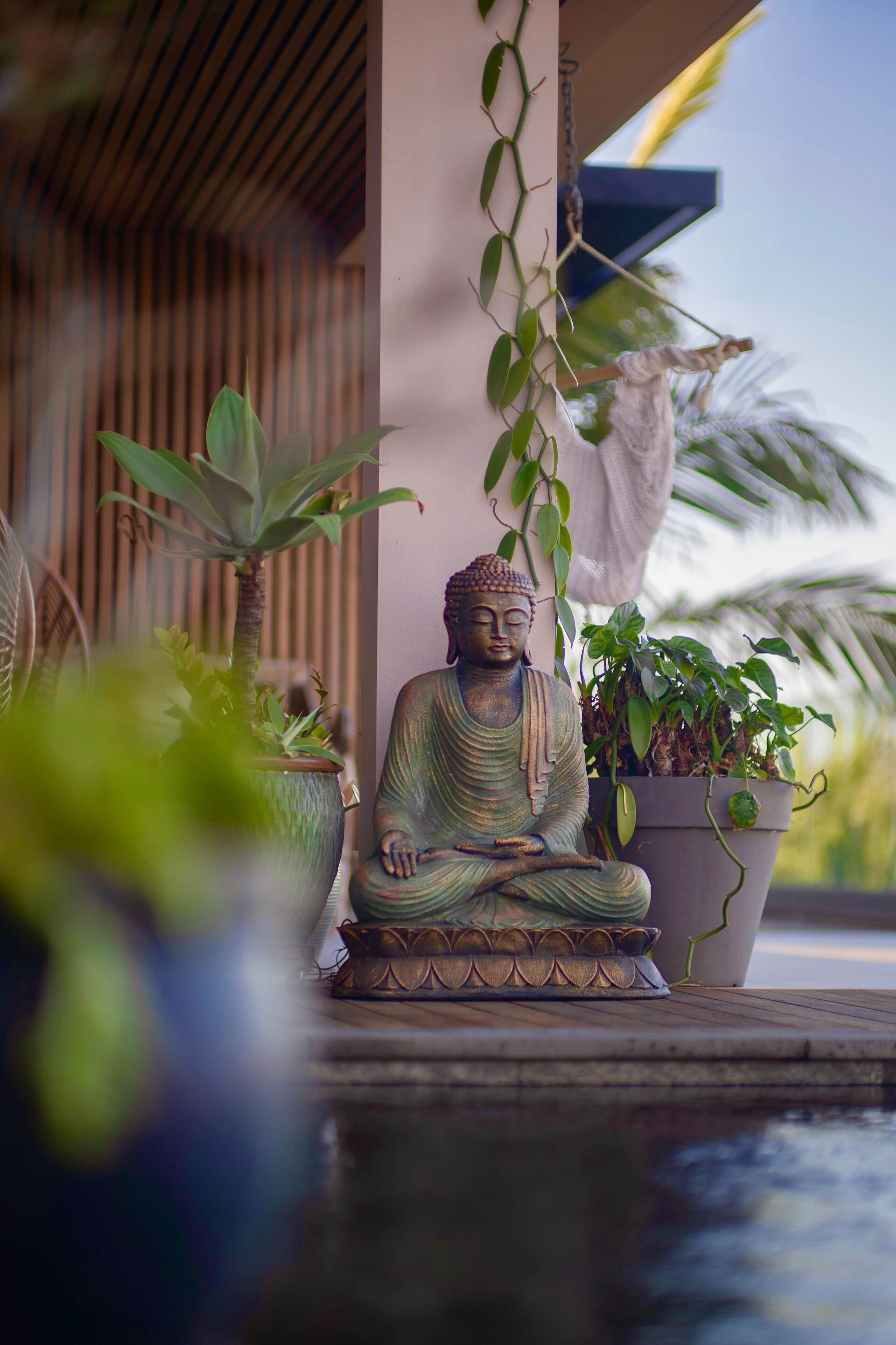 Eine Buddha-Statue in Meditationshaltung, umgeben von Topfpflanzen, mit einer Holzkonstruktion und einem Wasserspiel im Vordergrund.