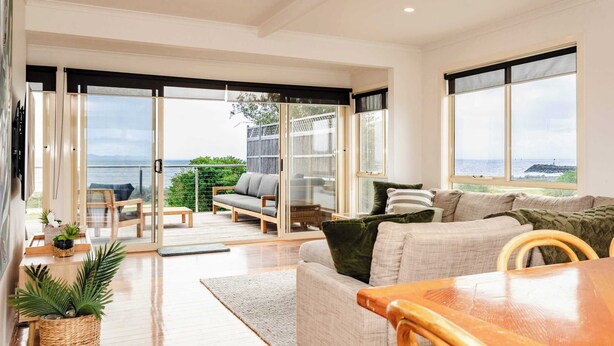 A spacious living area with a wooden dining table, a sofa, and a view of the sea through large windows.