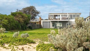A modern two-story house with a balcony, a fenced backyard, and a sandy area with vegetation.