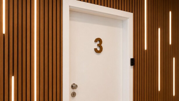 Una porta bianca con il numero 3, una serratura e una maniglia, incassata in una stanza con listelli di legno verticali e illuminazione a incasso.