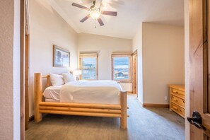 A bedroom with a wooden bed, a ceiling fan, and two windows with views of buildings.