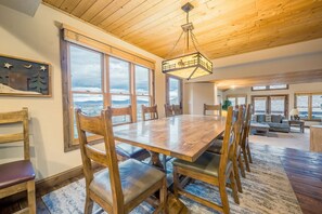 A spacious dining area with a large wooden table and chairs, large windows offering a view of the landscape, and a ceiling with wooden beams.