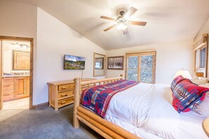 A bedroom with a wooden bed, a ceiling fan, a television, and a window with a view of trees.