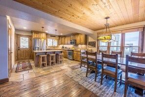 A spacious kitchen with wooden cabinets, a central island with stools, a dining table with chairs, and a large window providing a view of the outside.