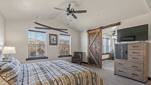 A bedroom with a large bed, a ceiling fan, two windows with blinds, a chair, a dresser, and a TV mounted on the wall. - Aspen Crest Condo, 4BR, Deer Valley, hot tub (Kamas)