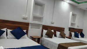 A hotel room with three single beds, each with a wooden headboard, white bedding, and blue and brown pillows. There are white nightstands, a ceiling fan, and built-in shelves above the beds.