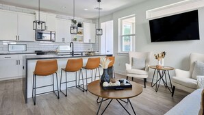 A modern kitchen with white cabinets, a central island with stools, a flat-screen TV, and a dining area with a round table and chairs.