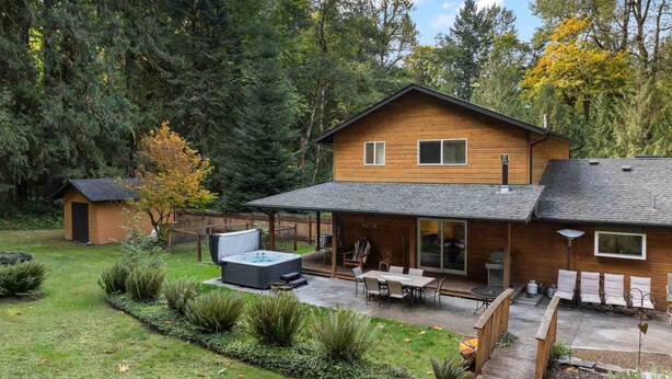 Una casa de madera con un patio, jacuzzi y zona de estar al aire libre, rodeada de un jardín y árboles.