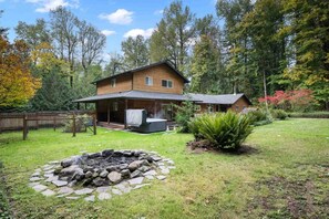 A two-story wooden house with a covered patio, a hot tub, and a stone fire pit in the yard. - Alpine Getaway w/ Hot Tub, Firepit & Stunning Views, Pet Friendly, Near Mt. Hood Slopes (Welches)