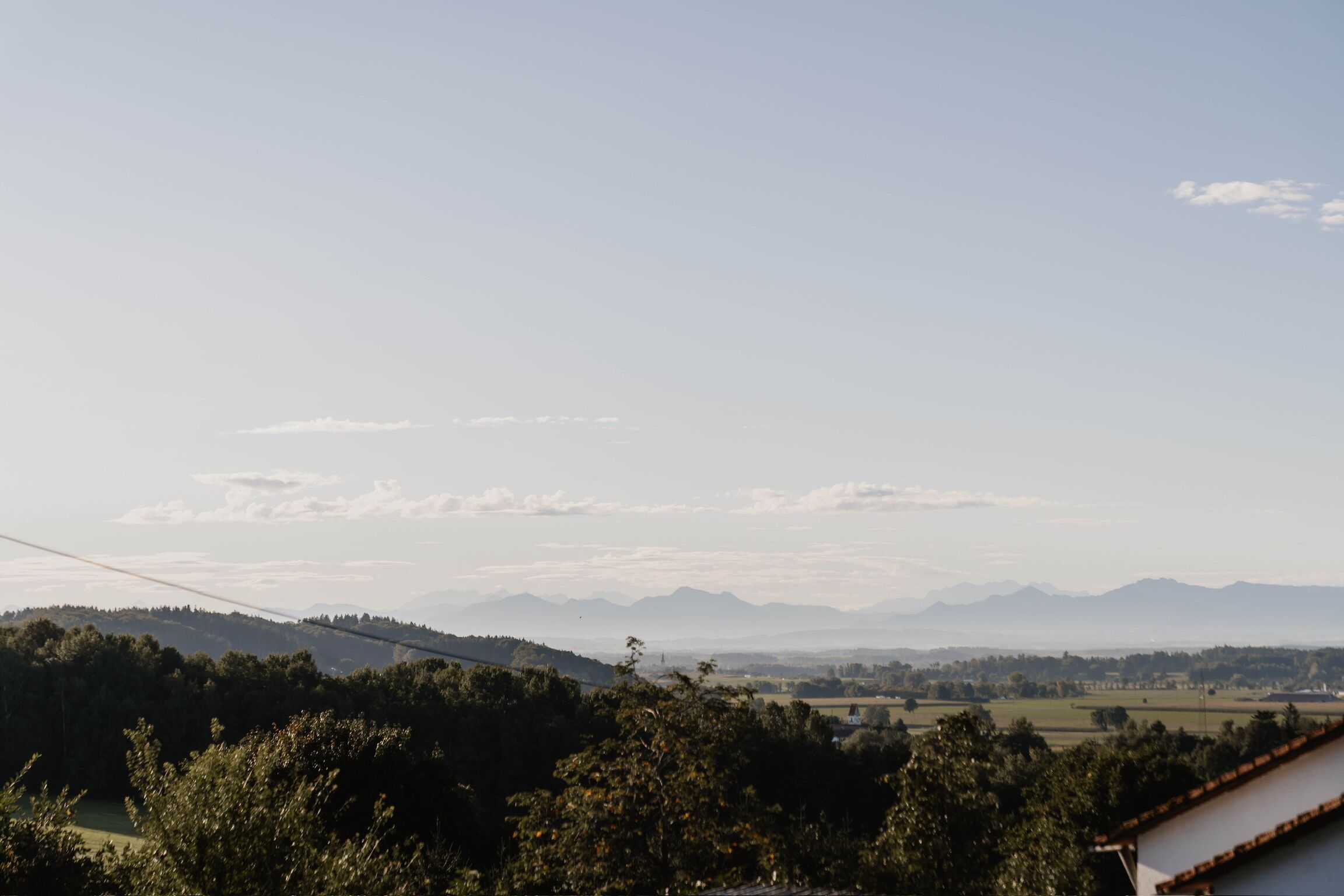 Ländliche Landschaft mit Blick auf ferne Berge, blauem Himmel und einem Haus mit rotem Dach im Vordergrund.