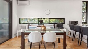 Un comedor moderno con mesa de vidrio, sillas blancas, una estatua de Buda como pieza central y vistas a través de ventanas amplias.