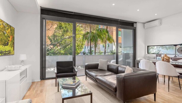 Una sala de estar moderna con sofá de cuero, una mesa de centro de vidrio y un ventanal con vistas a árboles y edificios.