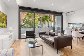 Un salon moderne avec un canapé en cuir, une table basse en verre et une grande fenêtre donnant sur des arbres et des immeubles.