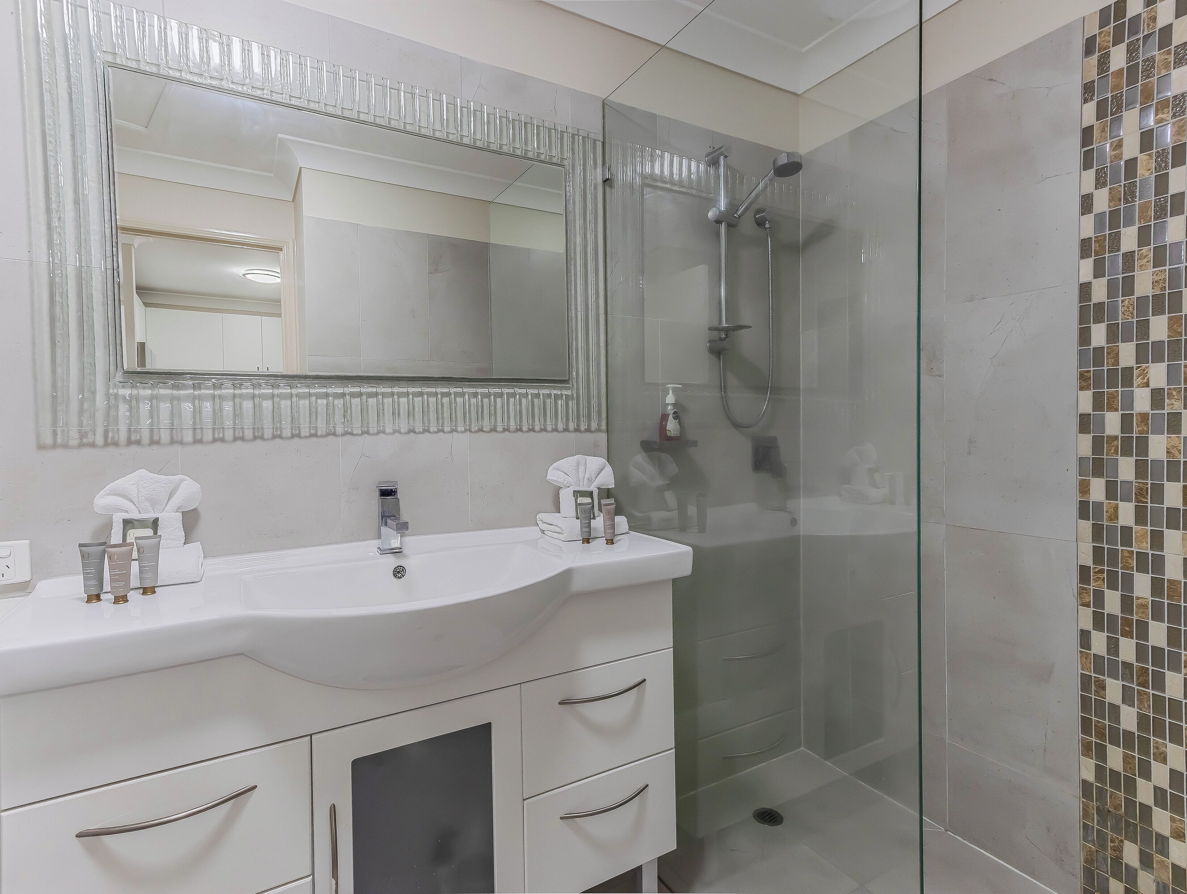 Un baño con cabina de ducha de vidrio, un lavabo blanco con dos grifos y un espejo grande sobre el lavabo.