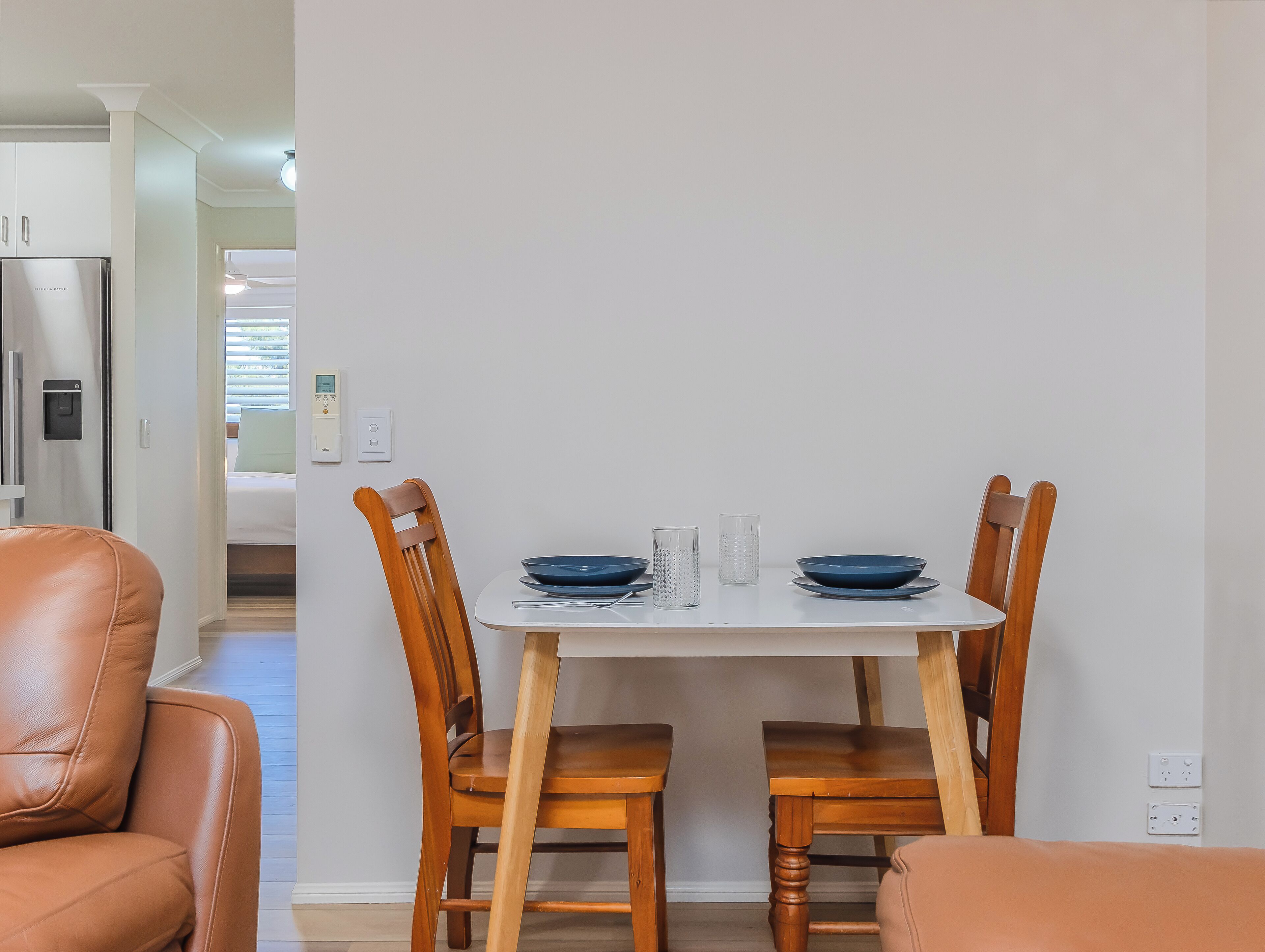 Un comedor con una mesa dispuesta para dos personas, dos sillas de madera, un mantel blanco y un refrigerador de acero inoxidable al fondo.