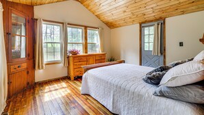 Un dormitorio con suelo de madera, una cama grande con ropa blanca, un aparador de madera y dos ventanas con cortinas.