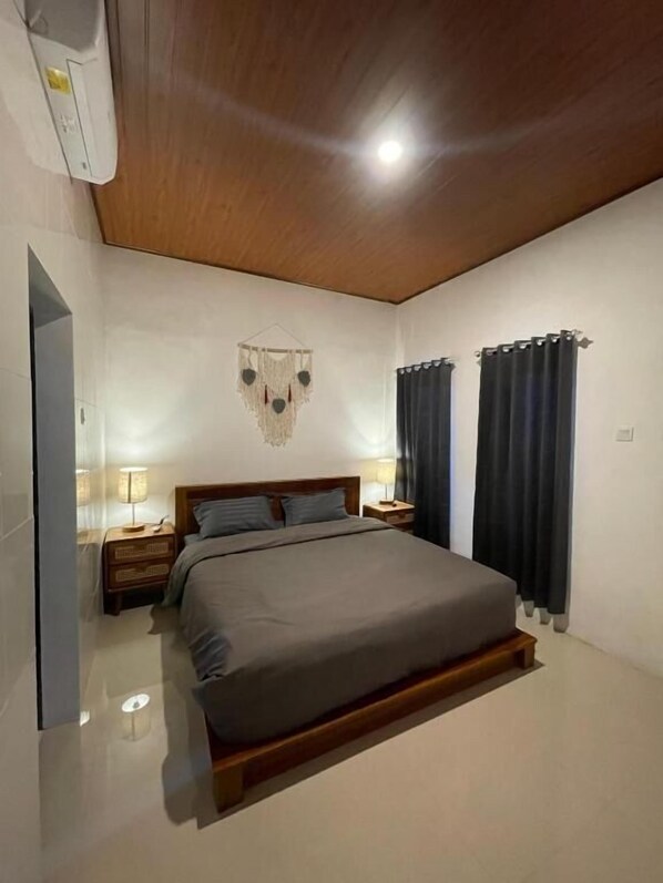A bedroom with a large bed, wooden headboard, two bedside tables with lamps, a wall hanging, and dark curtains.