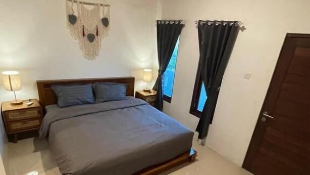 A bedroom with a large bed, two bedside tables with lamps, a wooden headboard, a wall hanging, and a door. - Pradnya Homestay - 1Bedroom Near Uluwatu (Ungasan)