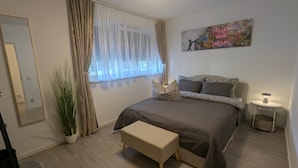A bedroom with a bed, a bedside table, a nightstand, a mirror, a window with curtains, and a wall art piece.