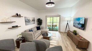 A modern living room with a sectional sofa, grey armchairs, a coffee table, and a TV mounted on the wall. There are shelves with decorative items and a painting on the wall.