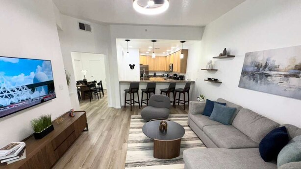 A modern living room with a sofa, coffee table, and TV. There is a kitchen area with bar stools and a dining table.