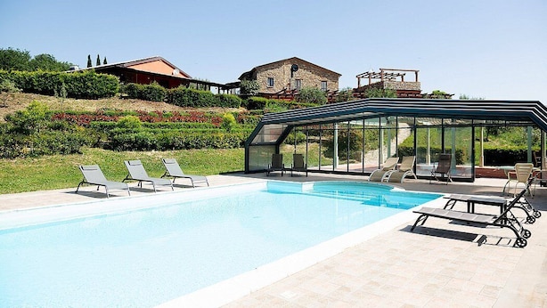 A swimming pool with lounge chairs, a glass-enclosed patio, and stone buildings in the background.