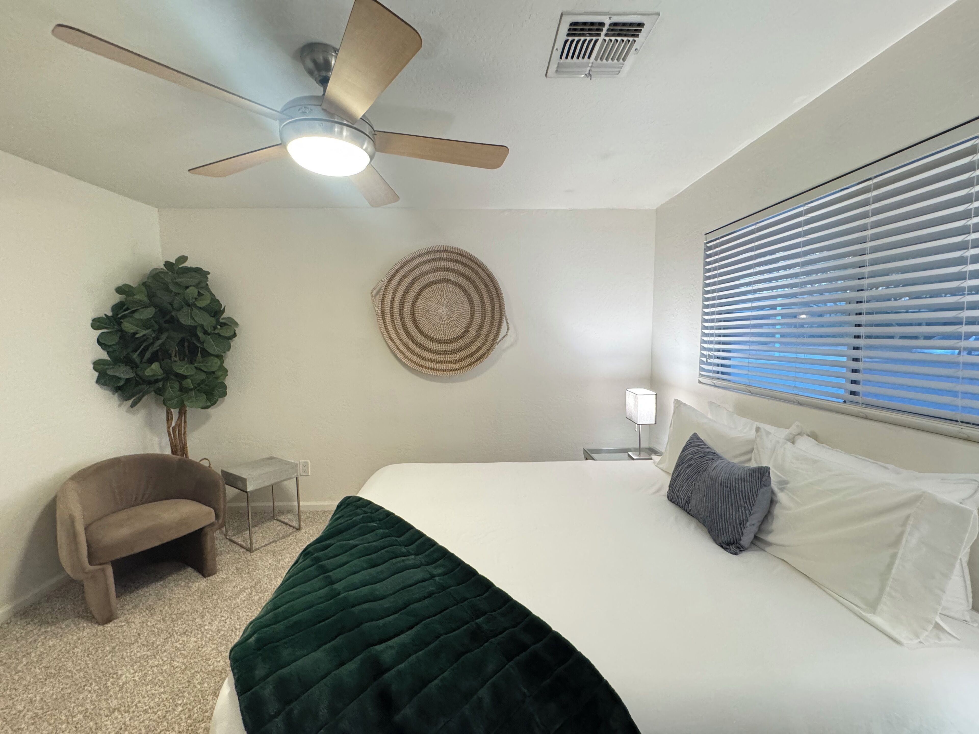 A bedroom with a bed, a ceiling fan, a wall-mounted air vent, a window with blinds, a small round rug, a potted plant, and a chair.