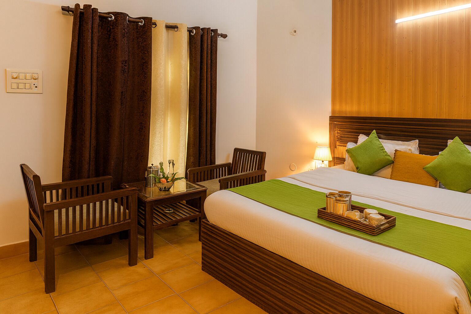 A hotel room with a bed, two wooden chairs, a small table with a plant, and a bedside table with a lamp.