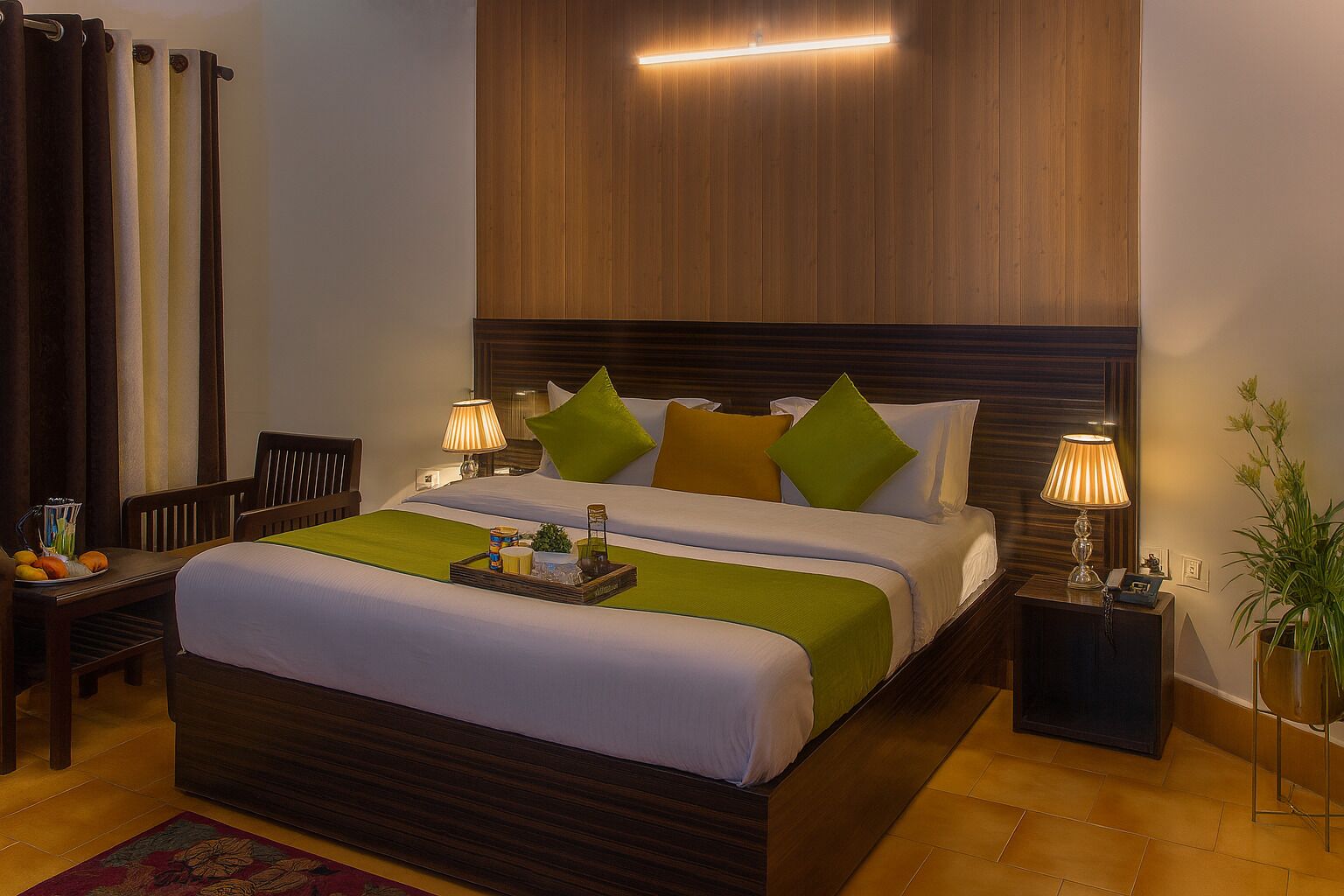 Habitación de hotel con una cama grande, dos mesitas de noche con lámparas, un cabecero de madera, una pequeña mesa con frutas y una bandeja, y una planta en maceta.