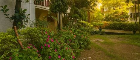 A garden with lush greenery and pink flowers, a pathway, and a building with a balcony.