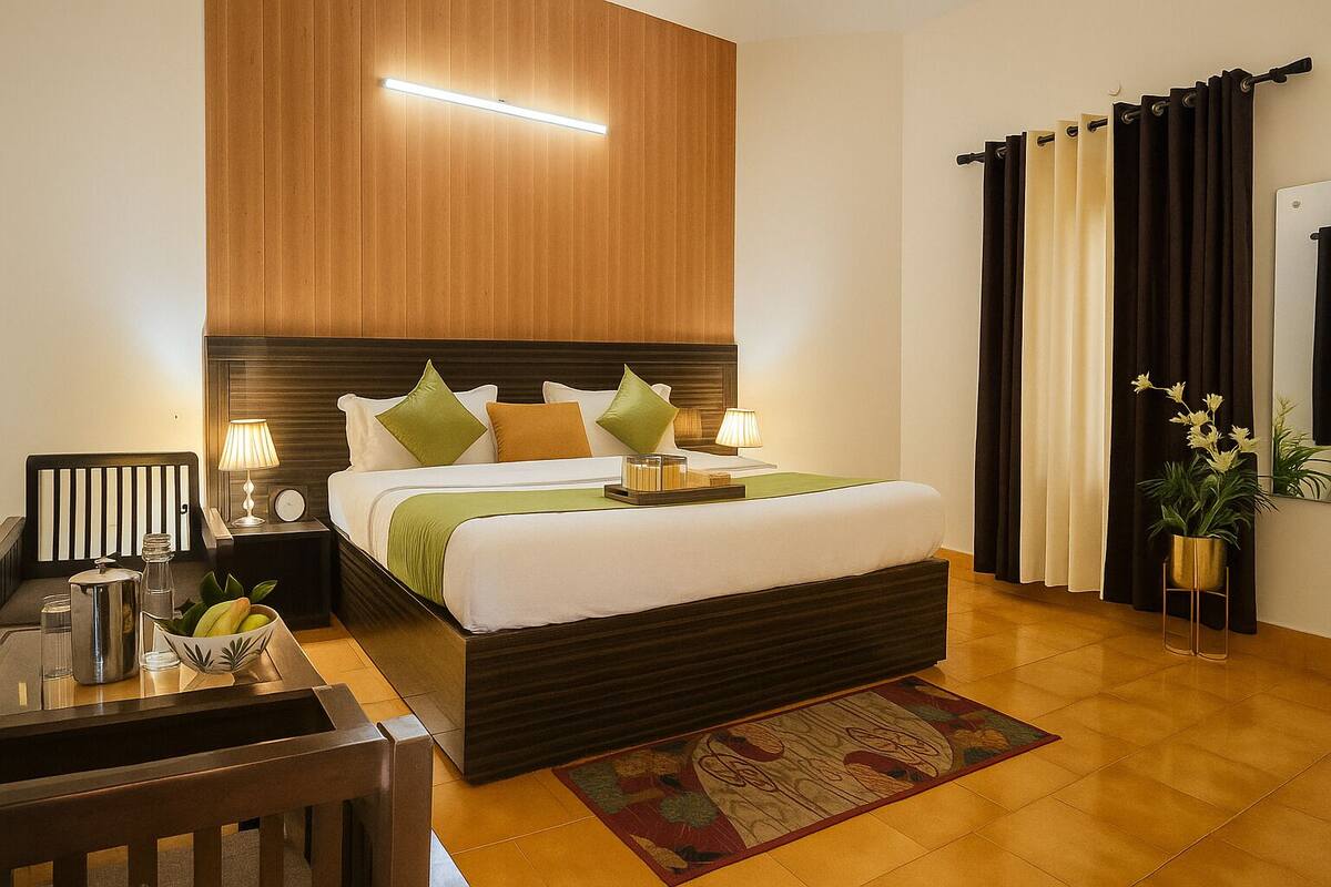 A hotel room with a wooden headboard, a bed with white bedding and green pillows, a bedside table with a lamp, a small table with a vase of flowers, and a window with curtains.