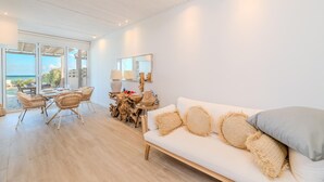 Un salon moderne avec un canapé blanc, des fauteuils en osier, une table à manger et une grande fenêtre donnant sur l’océan.