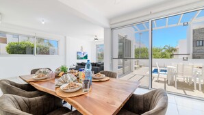 A dining area with a wooden table, four cushioned chairs, and a large glass sliding door leading to an outdoor patio with a pool and lounge chairs. - Elite Blu Heights Villa (Ayia Napa)