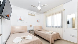 A hotel room with two beds, a ceiling fan, artwork on the walls, and a window with curtains. - Nissi Apartment #102 (Ayia Napa)